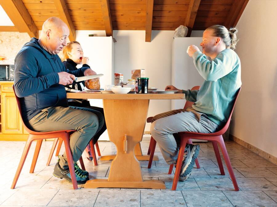 A dining area with a wooden table and chairs, a kitchen in the background, and a window providing natural light.