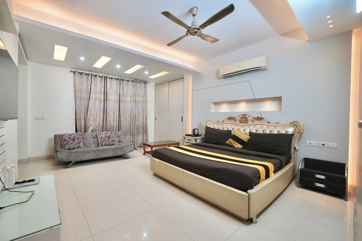 A bedroom with a bed, a sofa, a TV, a ceiling fan, and an air conditioning unit.