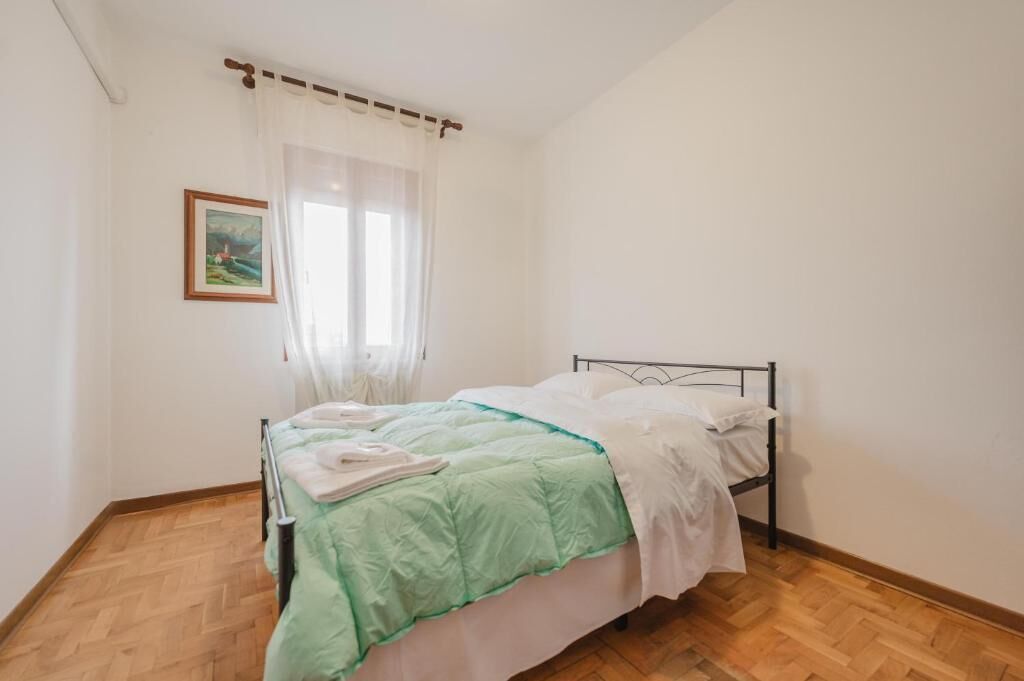 A single bed with a metal frame, white bedding, and a green blanket. There is a window with sheer curtains and a framed picture on the wall.