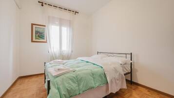 A single bed with a metal frame, white bedding, and a green blanket. There is a window with sheer curtains and a framed picture on the wall.