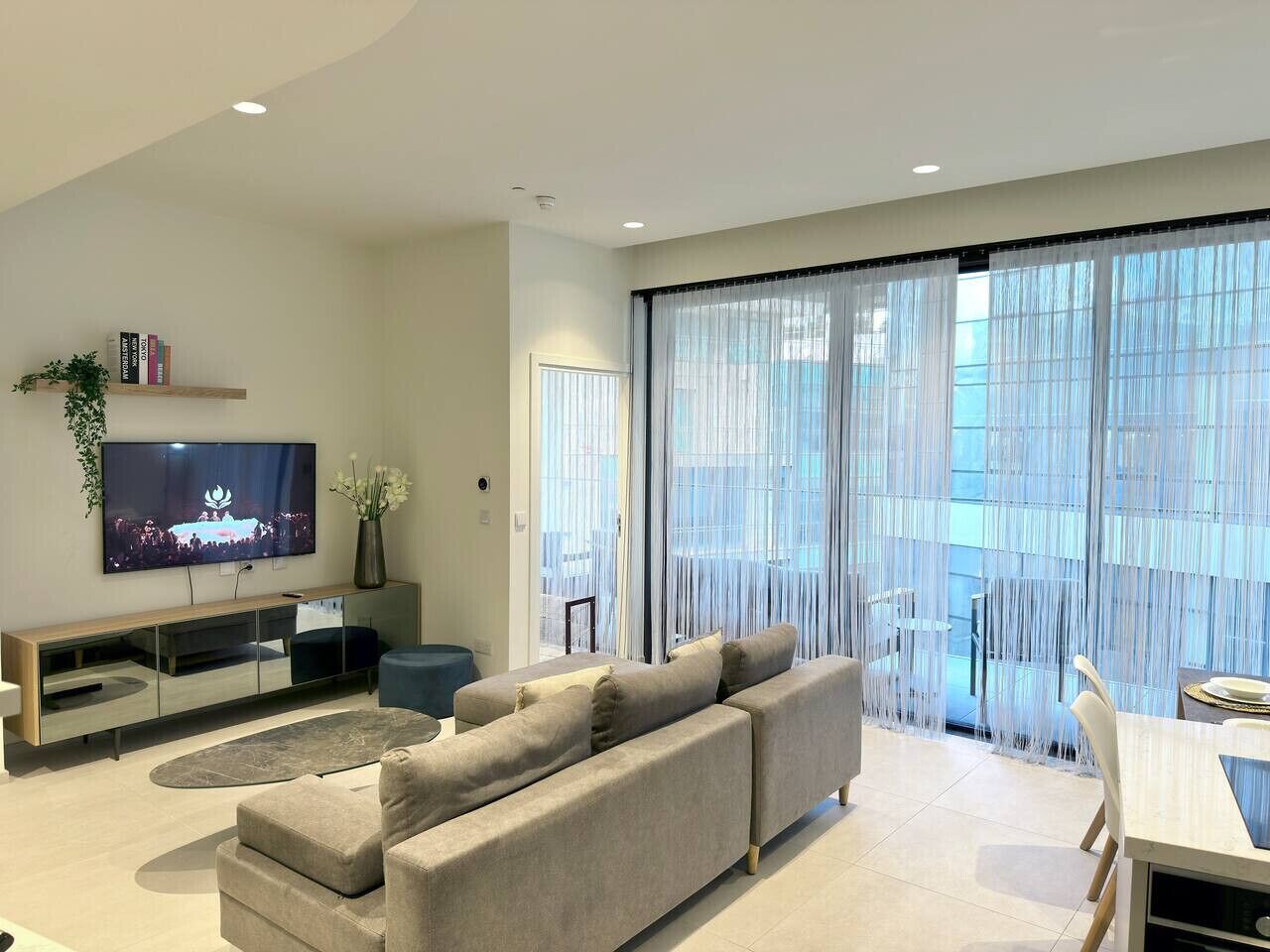 A modern living room with a flat-screen TV, a sofa, a dining table, and large windows with sheer curtains.