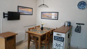 Quarto compacto com mesa de jantar de madeira, cadeiras, televisão presa na parede, quadros decorativos, relógio e placa indicando a localização do quarto.