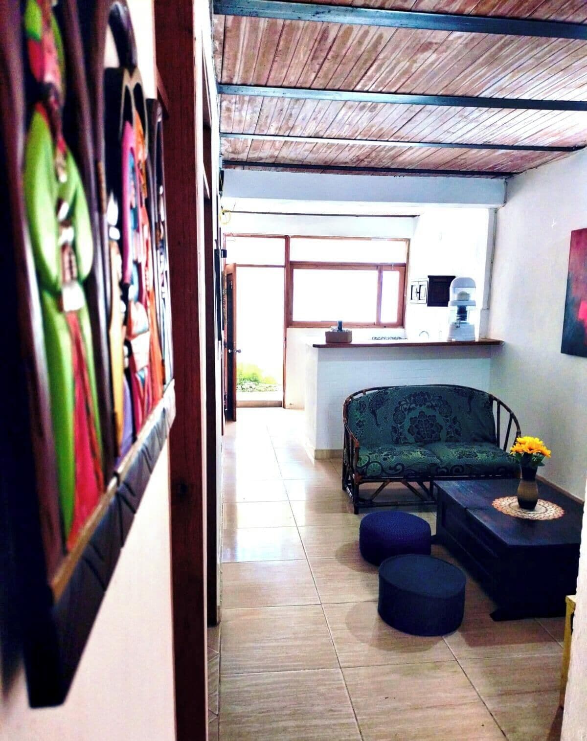 Una habitación con sofá, reposapiés y una mesita con un jarrón. Se ve una ventana y una puerta que da a otra habitación.