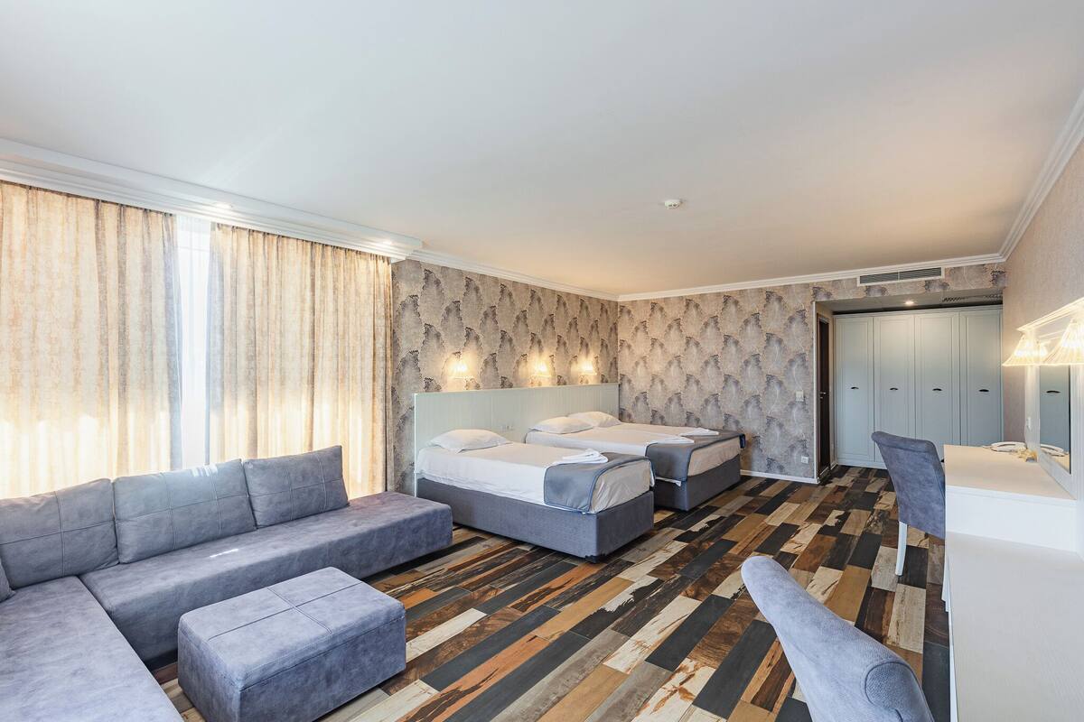 A modern hotel room with a sectional sofa, two single beds, a desk, and a wardrobe.