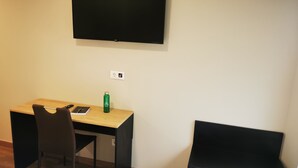 Una habitación con un escritorio, una silla, un televisor en la pared y una pequeña botella verde sobre el escritorio.