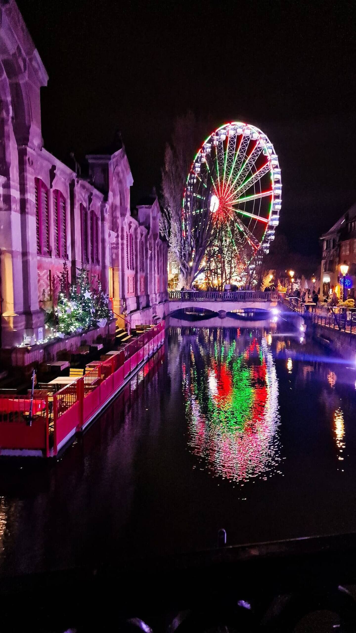 A nighttime view of a historic building with a large Ferris wheel in the background, reflecting on a calm water body.