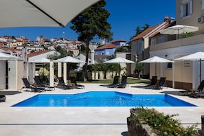 A swimming pool with lounge chairs and umbrellas, surrounded by buildings with red-tiled roofs.