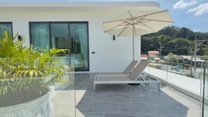 A modern balcony with a glass enclosure, a lounge chair, an umbrella, and a potted plant. - Phuvista Villas Naithon - Escape Villas  (Sa Khu)