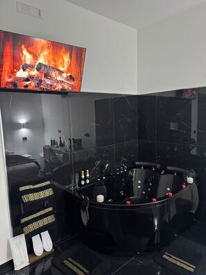 Ein modernes Badezimmer mit einer schwarzen Jaccuzzi-Badewanne, einem großen Spiegel und einem Fernseher, der einen Kamin zeigt.
