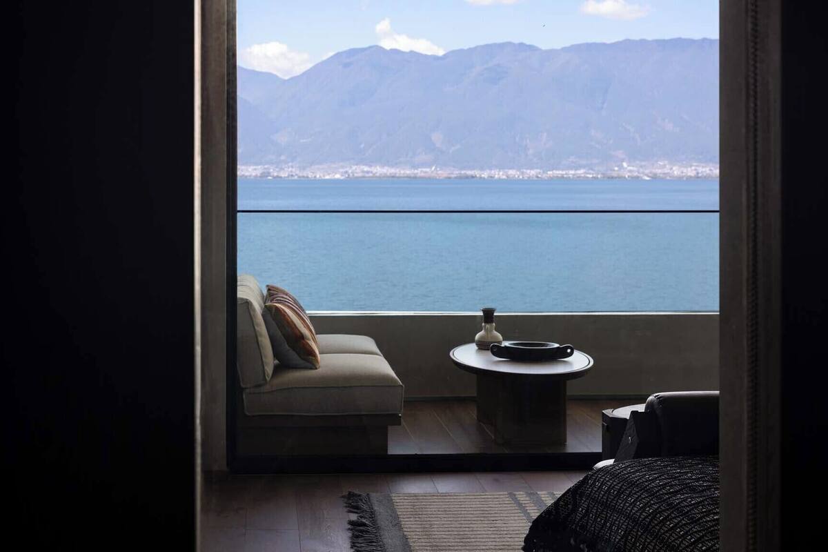 A room with a sofa and a small table, a view of a body of water and mountains, and a balcony door.