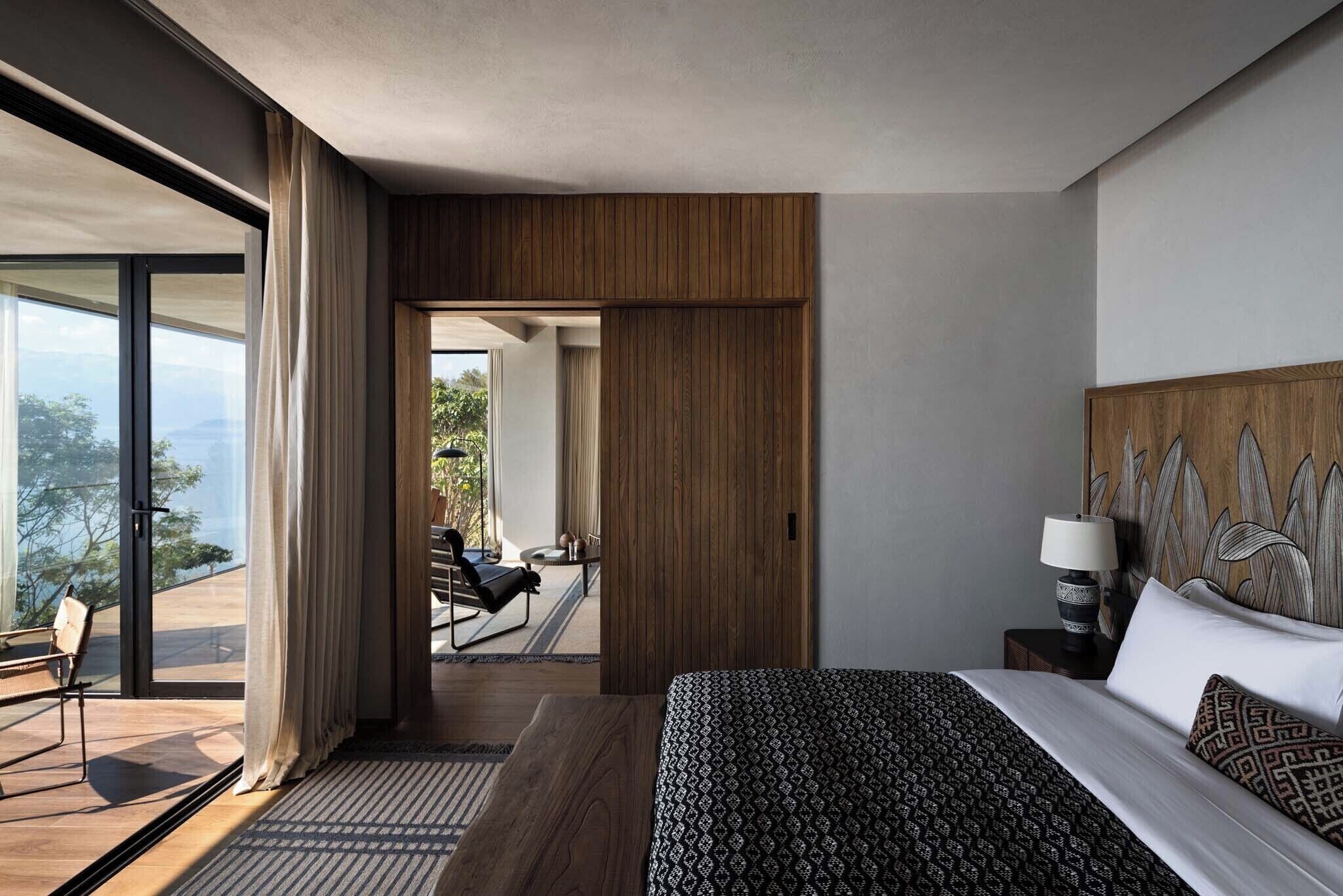 A modern hotel room with a large bed, a nightstand, a lamp, and a view of the outdoors.