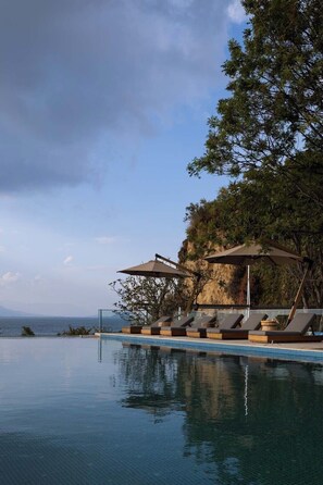 Área à beira da piscina com espreguiçadeiras e guarda-sóis, com vista para um corpo d'água e uma paisagem montanhosa.