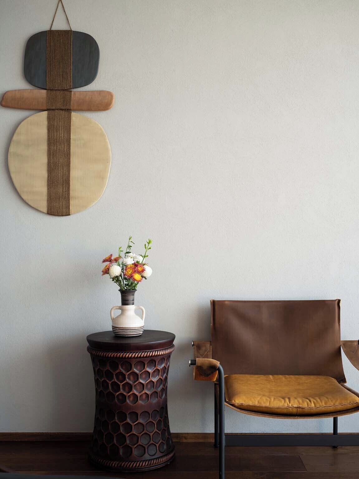 Una habitación con una mesita redonda de madera, un florero con flores, un sillón de cuero marrón y una pared decorada con arte geométrico.