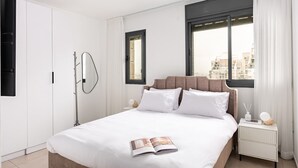 A modern hotel room with a large bed, a bedside table with a lamp, a window with curtains, and a view of buildings outside.