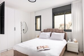 Ein modernes Hotelzimmer mit einem großen Bett, einem Nachttisch mit Lampe, einem Fenster mit Vorhängen und Blick auf Gebäude.