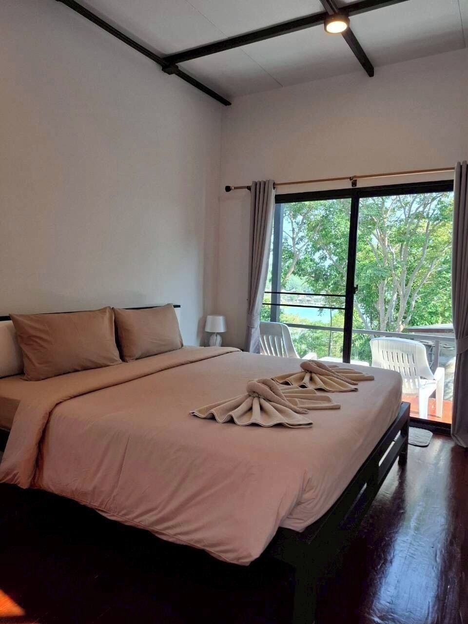 A bedroom with a large bed, a window with curtains, a balcony with chairs, and a ceiling with exposed beams.