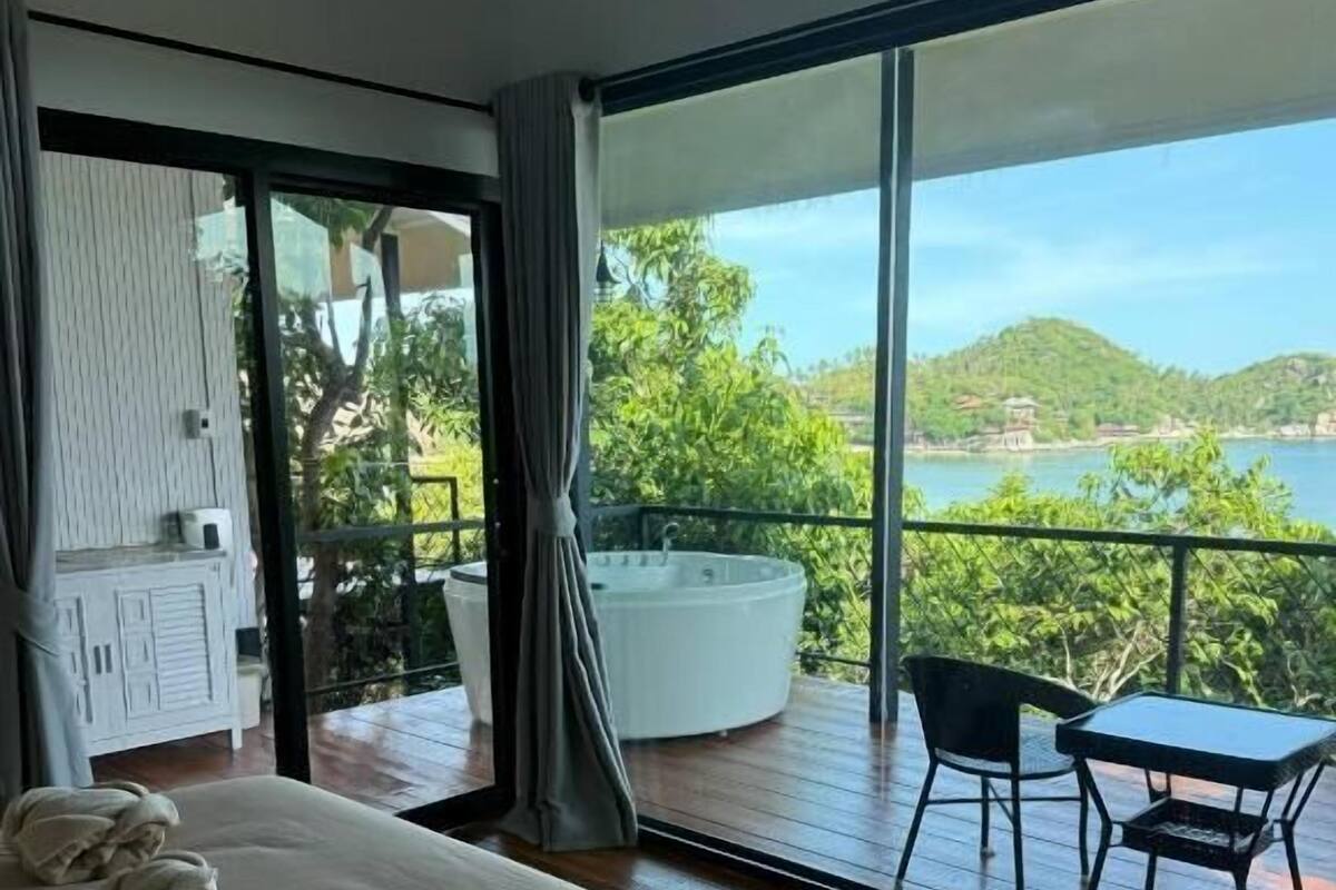 Habitación de hotel con una cama grande, un balcón con mesa y sillas, y vistas a un cuerpo de agua y vegetación.