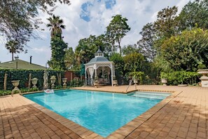 A swimming pool with a gazebo, surrounded by lush greenery and a brick patio.