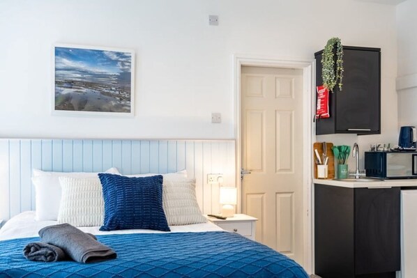 Habitación de hotel con cama, mesita de noche, televisor de pared, puerta y una pequeña cocina con microondas.