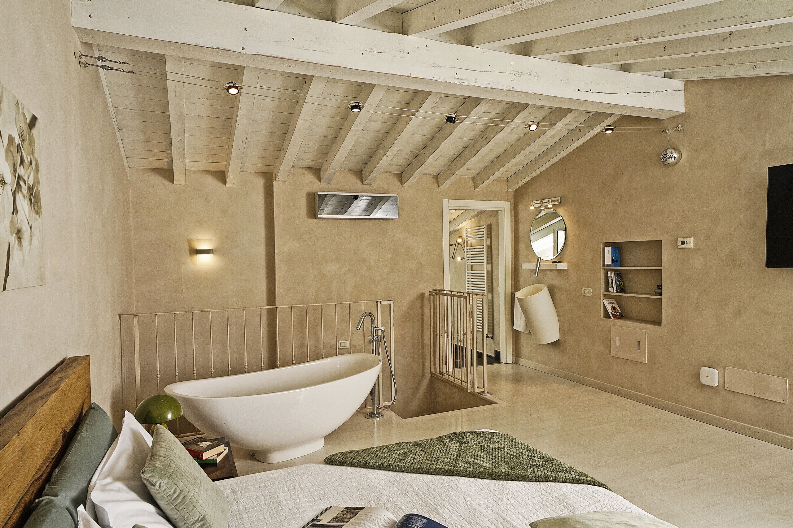 A bedroom with a large bathtub, a bed with pillows, a bedside table, and a mirror.