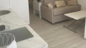 Uma cozinha moderna com bancada de mármore, uma sala de jantar com sofá e mesa pequena, e uma área de estar com sofá e planta.