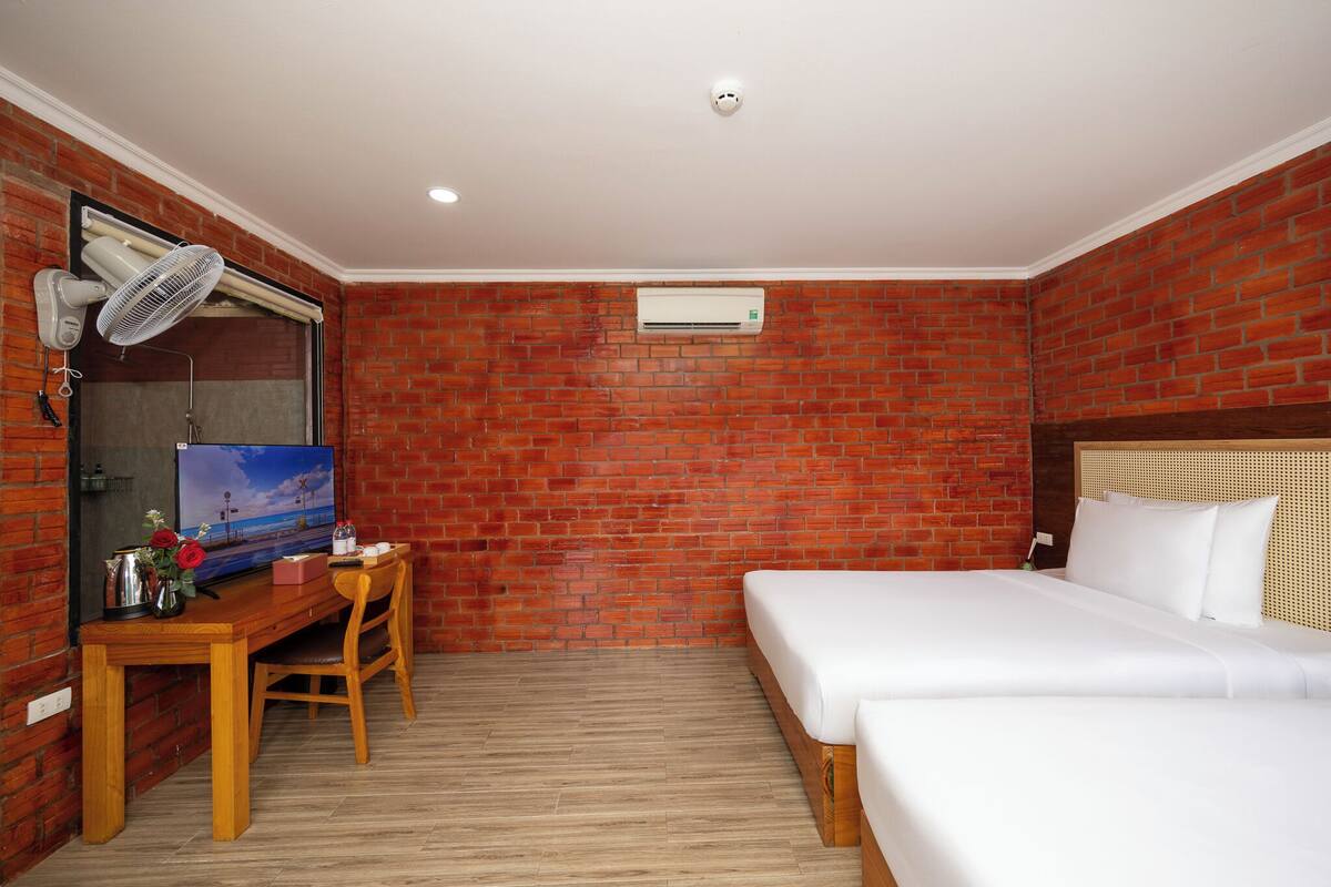 A hotel room with a brick wall, a wooden desk with a TV, a bed, and an air conditioning unit.