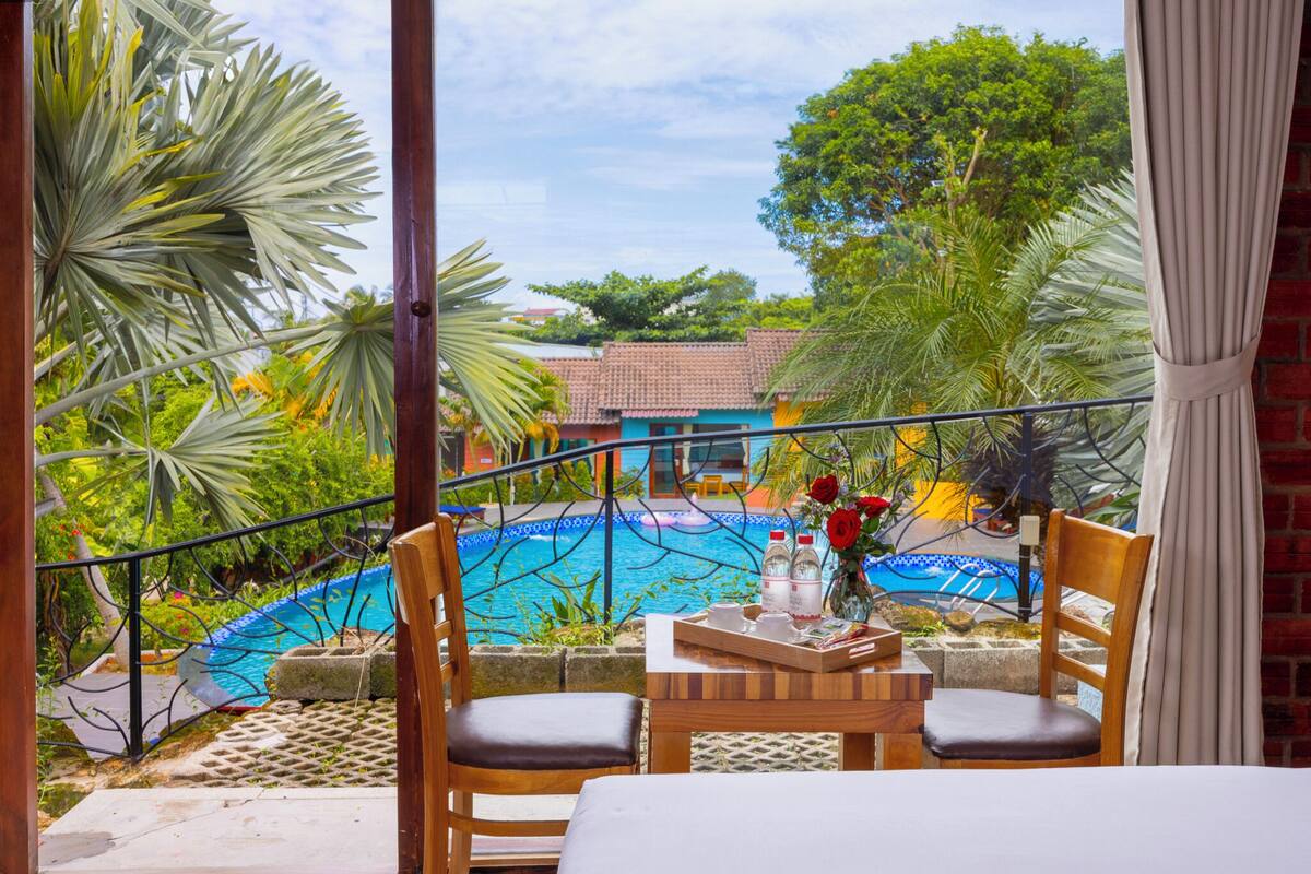A balcony with a view of a swimming pool, surrounded by tropical plants and a building with a tiled roof.