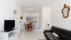 Ein modernes Wohnzimmer mit einer schwarzen Ledersofa, einem Flachbildfernseher auf einem weißen Standfuß, einem Essbereich mit Tisch und Stühlen sowie einer Küche im Hintergrund.
