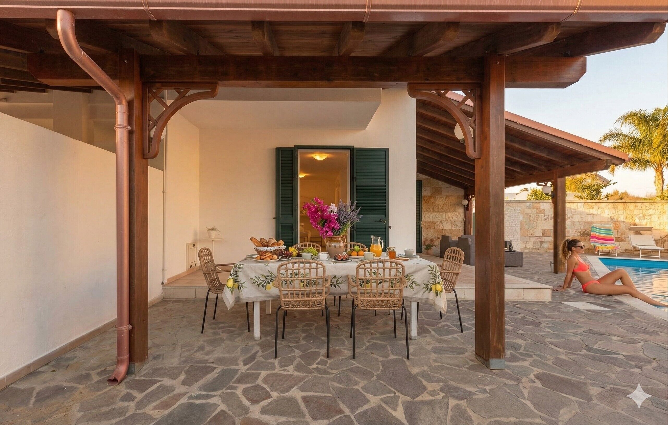 An outdoor dining area with stone flooring, a table set with food and drinks, and a pool in the background.