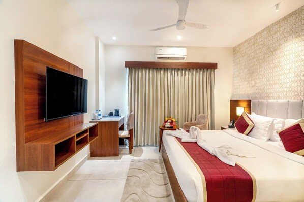 A hotel room with a flat-screen TV, a desk with a chair, a bed with a red bedspread, and a ceiling fan. - Passions Hotel (Kuttanad Taluk)