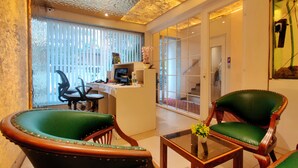 Een kamer met een bureau, twee groene leren fauteuils, een glazen salontafel en uitzicht door een raam met jaloezieën.