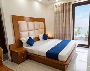 Habitación de hotel con cabecera de madera, cama con ropa de cama blanca y azul, mesita de noche de madera, un cajón y vistas al exterior a través de una ventana.
