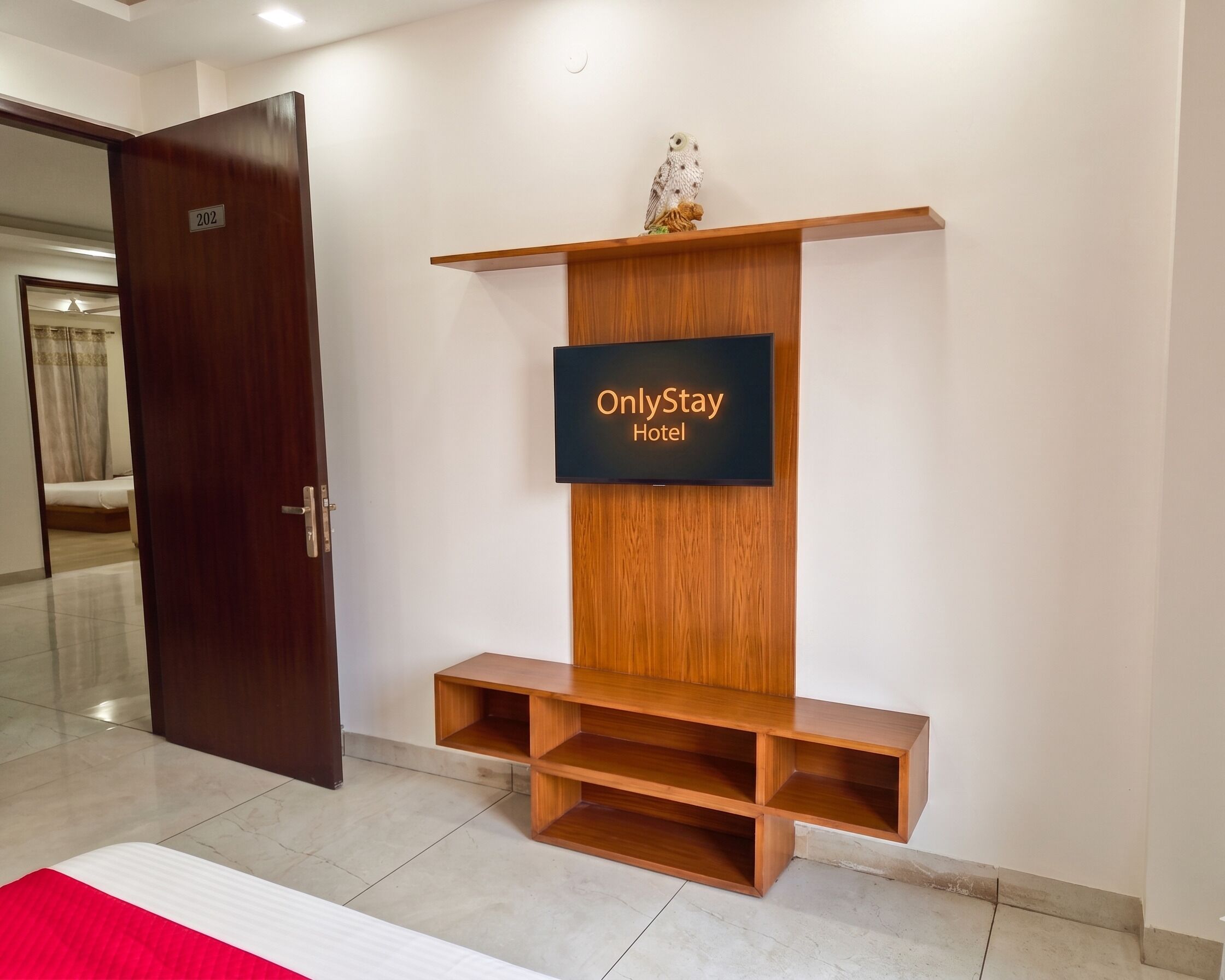 Un hall d'hôtel avec un comptoir d'accueil en bois, un écran numérique affichant l'enseigne « OnlyStay Hotel », une porte avec une plaque numérotée, et un aperçu d'une chambre avec un lit.