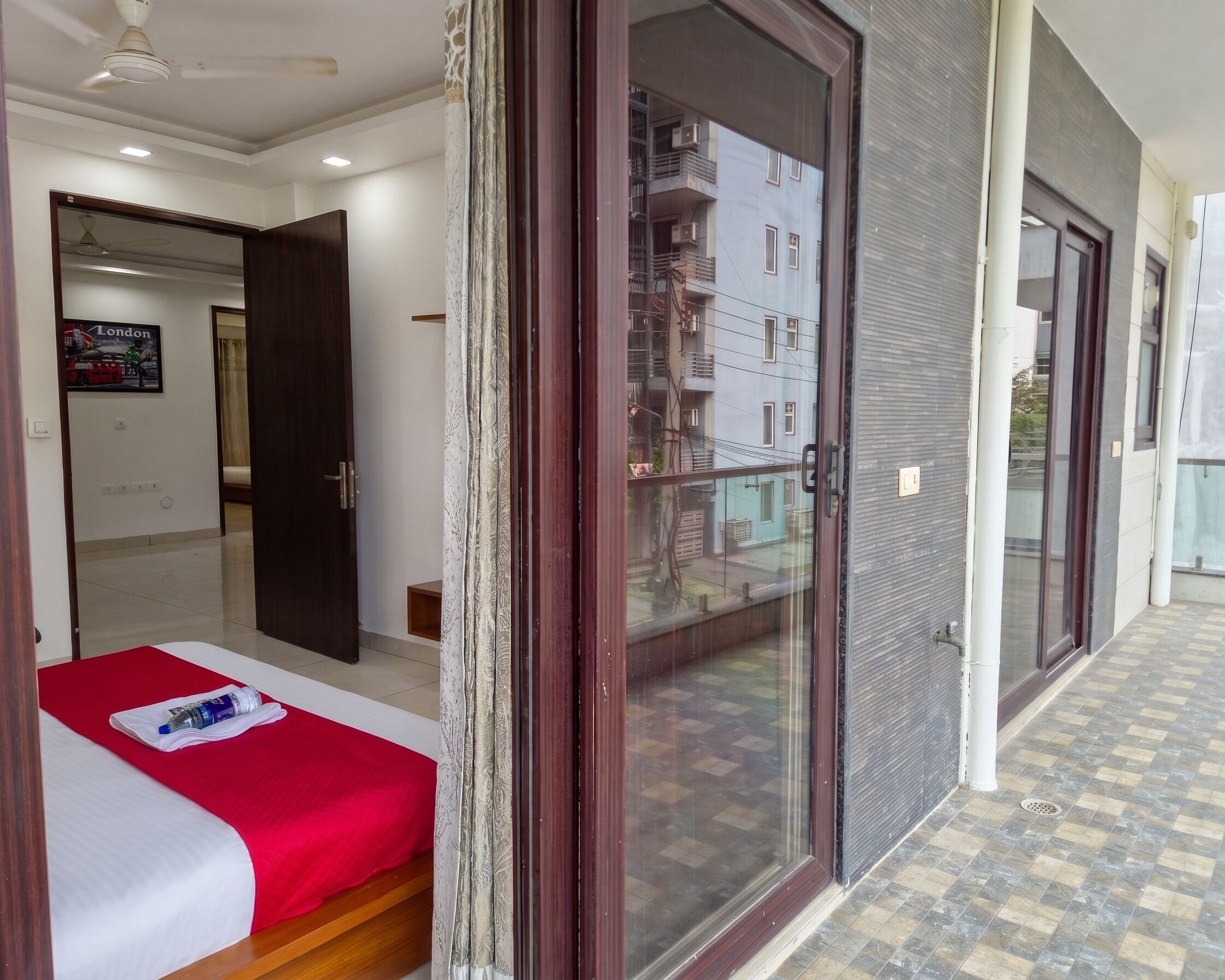 A hotel room with a bed, a red and white bedspread, a water bottle, a door leading to a balcony, and a view of a building with multiple windows.