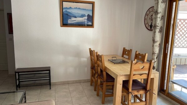 A dining area with a wooden table and chairs, a small bench, a framed picture on the wall, and a view through a glass door.