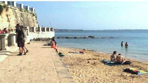 Una playa con gente tomando el sol y nadando, un muro de piedra y un cielo despejado.