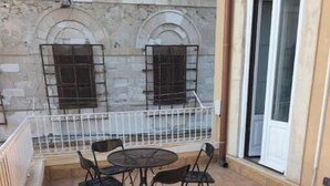 Un patio con una mesa redonda y sillas, baranda blanca, y un edificio de paredes de piedra y ventanas.