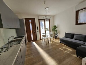 Eine moderne Küche mit eingebautem Herd, ein Essbereich mit Tisch und Stühlen und ein dunkelgraues Sofa im Wohnbereich.