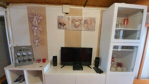 Ein Zimmer mit Fernseher, Lautsprechern, einem kleinen Tisch und einer Schranke mit Glastüren.