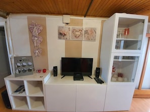 Ein Zimmer mit Fernseher, Lautsprechern, einem kleinen Tisch und einer Schranke mit Glastüren.