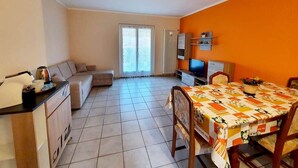 Una sala de estar con sofá, una mesa de comedor con sillas, televisor y un área de cocina con armarios y un radiador.