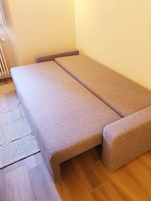 A compact bed with a grey mattress and matching headboard and footboard, positioned against a wall with a radiator and a wooden floor. - ELEONORA APARTMENT A FEW METERS FROM THE LAKE (Verbania)