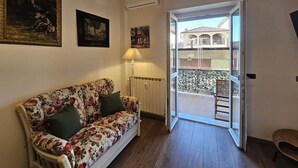 Ein Zimmer mit einer blumigen Couch, einer Lampe, einem Heizkörper und einer Glastür, die zu einem Außenbereich mit einem Sessel führt.