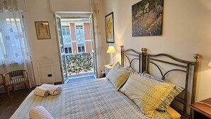 Ein Schlafzimmer mit einem Bett, einem Sessel, einem Nachttisch, einem Fenster mit Vorhängen und einem Bild an der Wand.