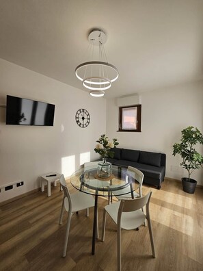 A modern living room with a round glass dining table, a black sofa, a potted plant, a mounted TV, and a wall clock. - GREY 2 (air conditioning and free private parking) (Verbania)