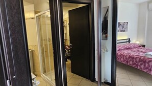 Una habitación con cama, mesita de noche, espejo y puerta que da a un baño con ducha.