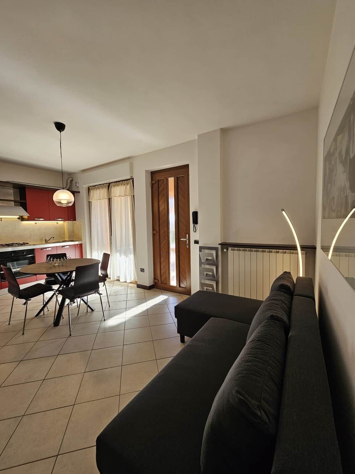 Un salon moderne avec un canapé, un coin repas avec des chaises, une cuisine avec des meubles rouges et une porte donnant sur l’extérieur.