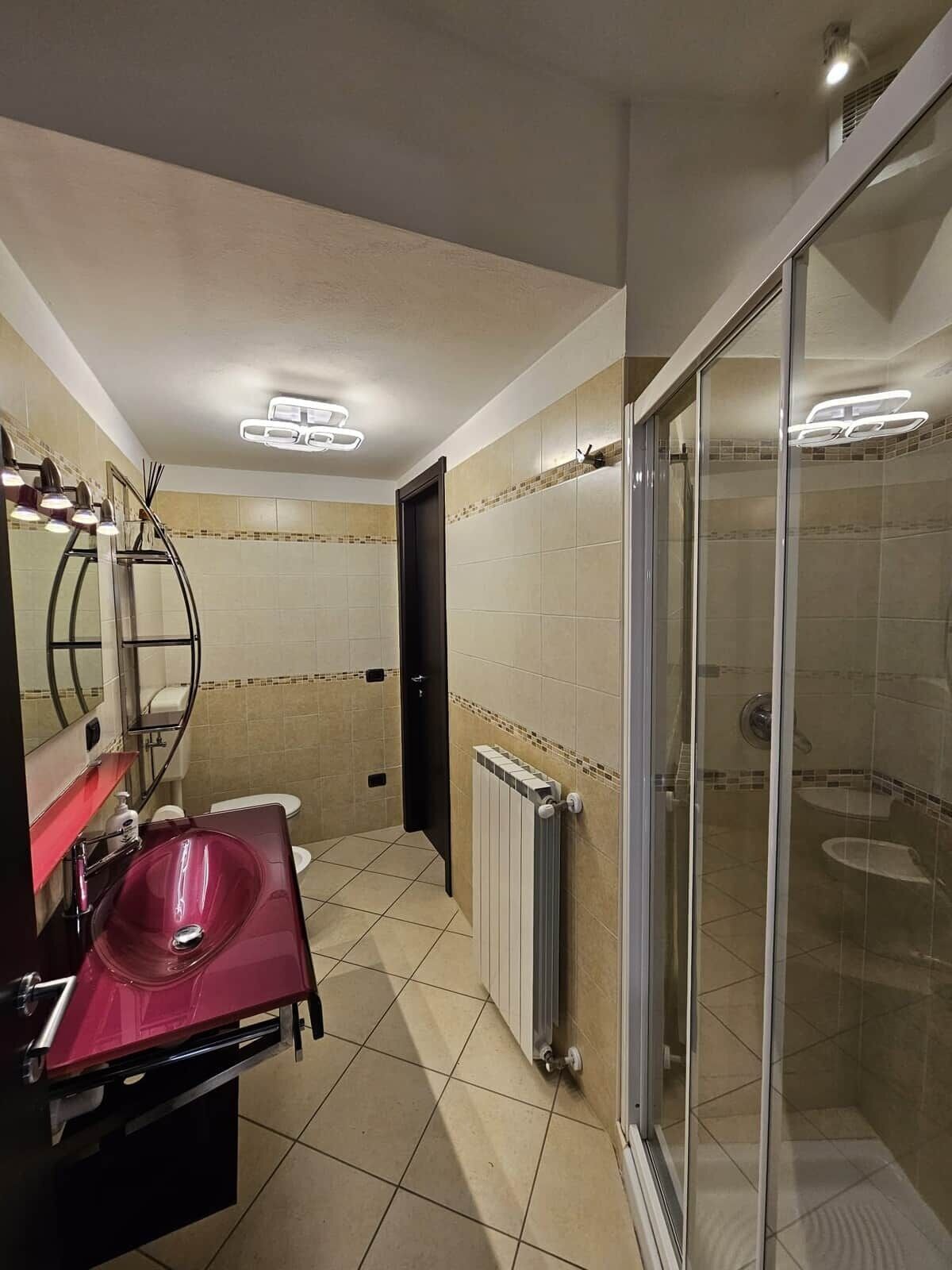 A bathroom with a glass shower enclosure, a vanity with a red sink, and a mirror with lights above.