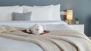 Una cama bien hecha con sábanas blancas, una almohada estampada, una mesita de noche con una lámpara y una bandeja decorativa con un hervidor.