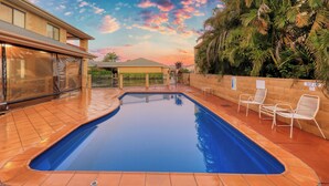 Una piscina con suelo de baldosas, un área de estar cubierta y vista a un edificio con fachada de ladrillo.